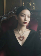 Asian woman looking off to the side wearing a hand-crafted red coral, heart-shaped stainless steel pendant necklace on a dark fabric background in a gothic setting.